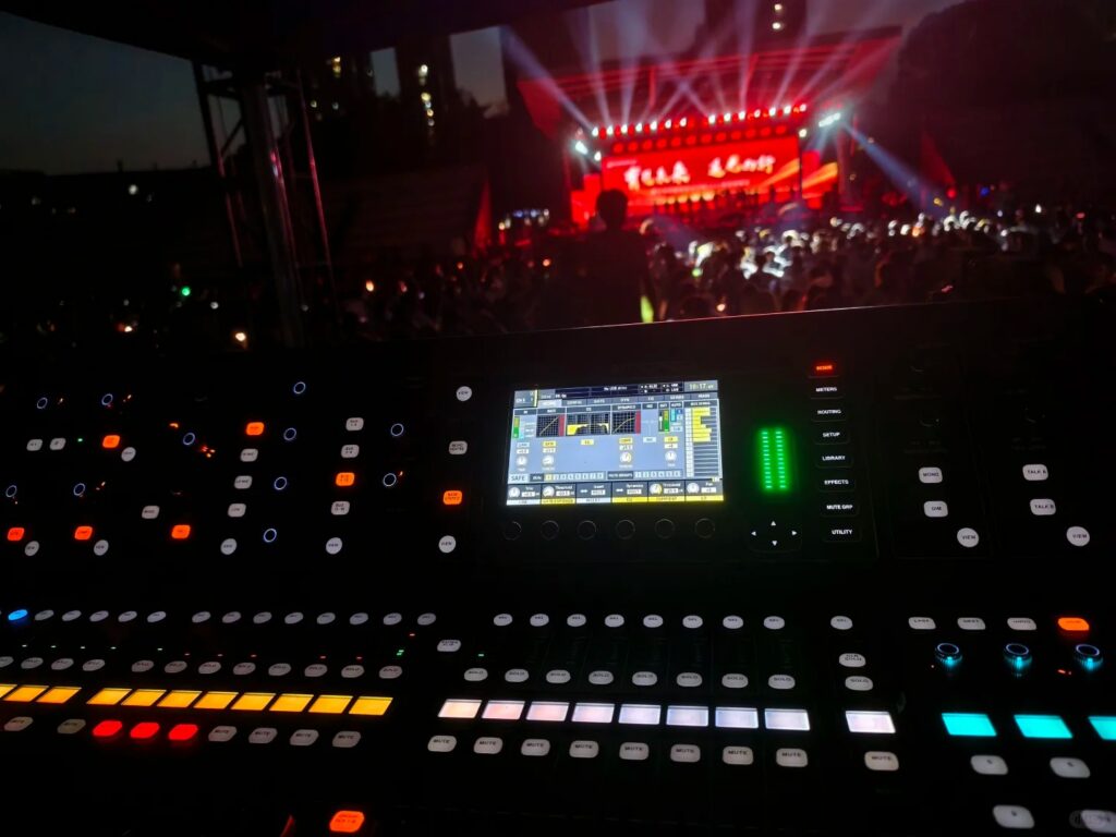 Interface de console de mixage numérique Allen & Heath éclairée en régie FOH lors d'un concert en plein air de nuit.